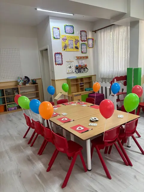 Balonlarla süslenmiş masa ve kırmızı sandalyelerle aydınlık sınıf